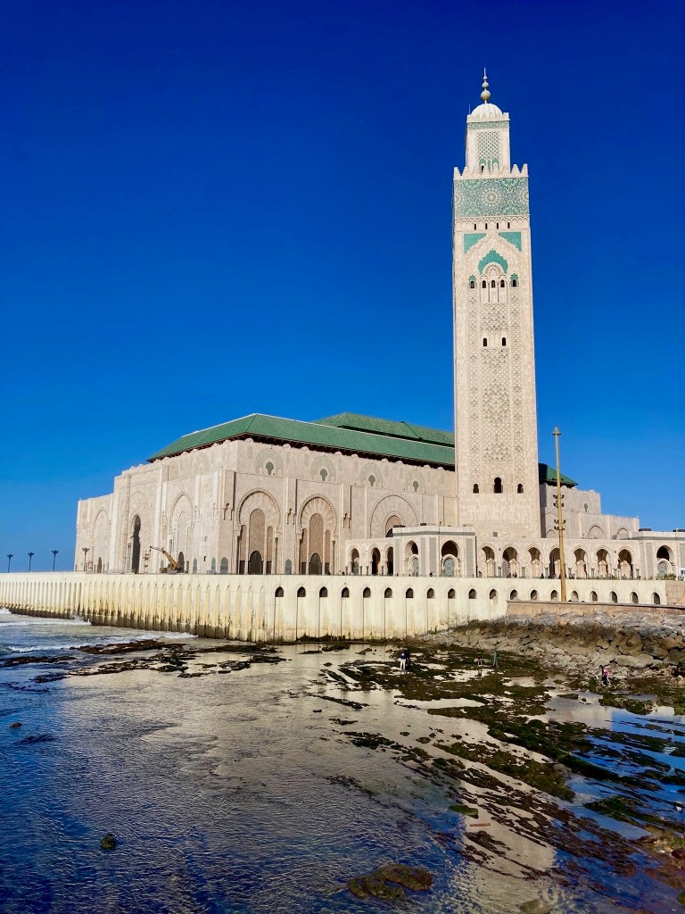 photo of a large white building with green tile roof and a towering minaret decorated with green tile, perched on the edge of water with a blue sky above
