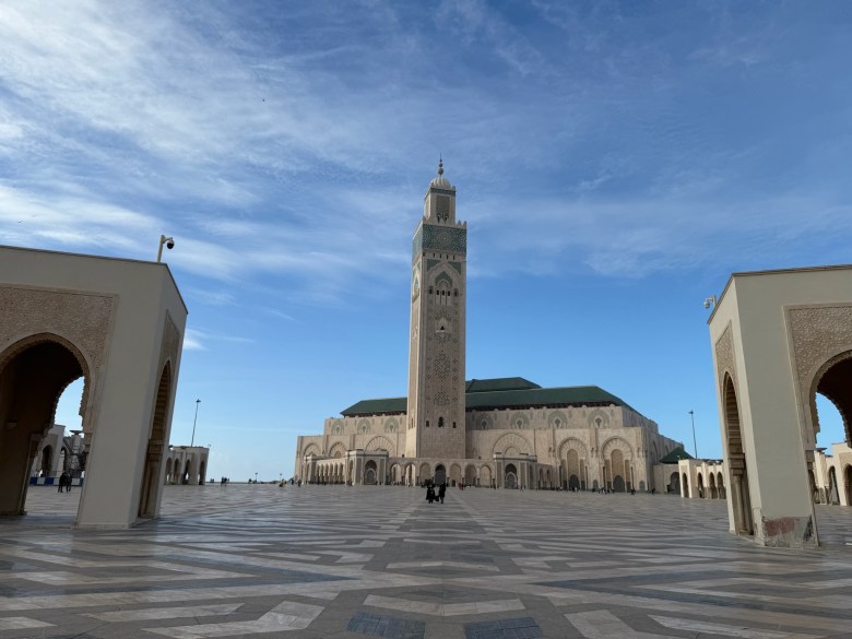 photo of a gray, brown, and green decorated rectangular tower in front of a lower rectangular building with a green tiled roof, set back across a wide expanse of gray and brown tiles and framed by single story arches