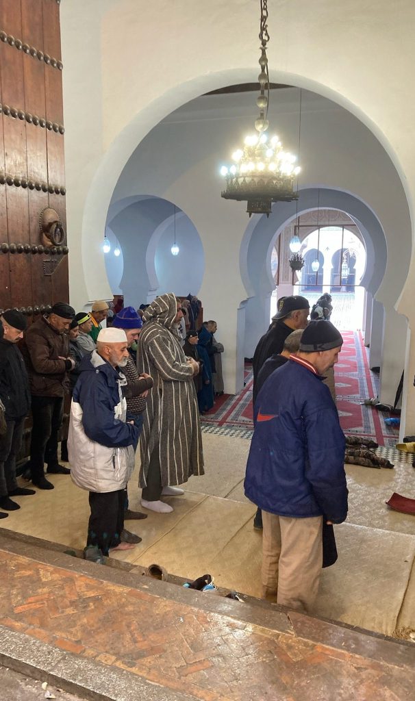 photo of men standing in socks in front of a series of arches in white walls, all facing to the right with heads down and hands clasped in front of them