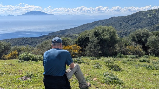 photo of a man in a blue shirt sitting on a grassy green bluff looking across water with low clouds and a mountain in the distance