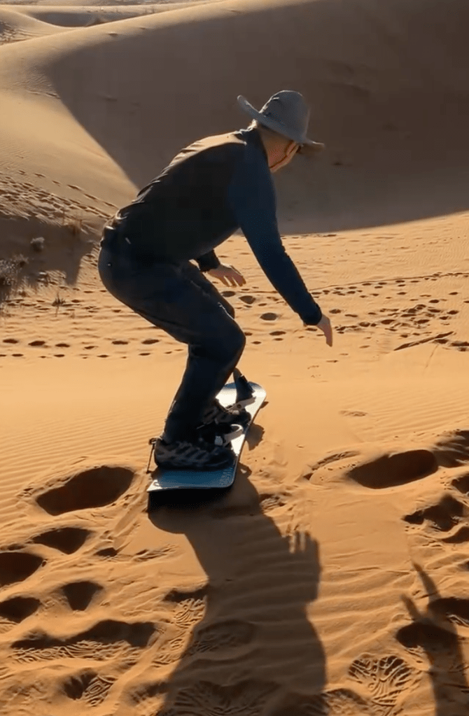 photo of a man wearing gray and a wide-brimmed sun had, crouched on a snowboard pointing down a golden brown sand dune