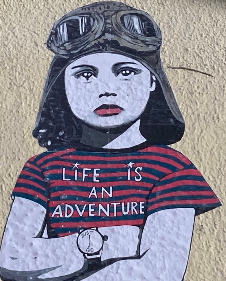 photo of a street art painting on a stucco wall depicting a child wearing an old-fashioned, great, goggled pilot's hatand a blue and red striped T-shirt that reads Life Is an Adventure. Her arms are crossed and she wears a watch that reads Just Go.