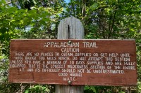 photo of a brown wood sign on a post against a backdrop of green leafy trees. The sign says APPALACHIAN TRAIL - CAUTION. There are no places to obtain supplies or get help until Abol Bridge 100 miles north. Do not attempt this section unless you have a minimum of 10 days supplies and are fully equipped. This is the longest wilderness section of the entire AT and its difficulty should not be underestimated. Good Hiking! M.A.T.C.