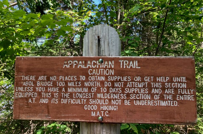 photo of a brown wood sign on a post against a backdrop of green leafy trees. The sign says APPALACHIAN TRAIL - CAUTION. There are no places to obtain supplies or get help until Abol Bridge 100 miles north. Do not attempt this section unless you have a minimum of 10 days supplies and are fully equipped. This is the longest wilderness section of the entire AT and its difficulty should not be underestimated. Good Hiking! M.A.T.C.
