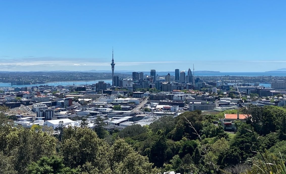 wide photo of a city skyline featuring a needle tower and several skyscrapers agains a blue sky and water, with green leafy trees in the foreground.
