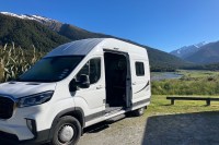 photo of a white camper van with the side sliding door open, backed into a campsite with a long green valley behind it and snow capped peaks in the distande