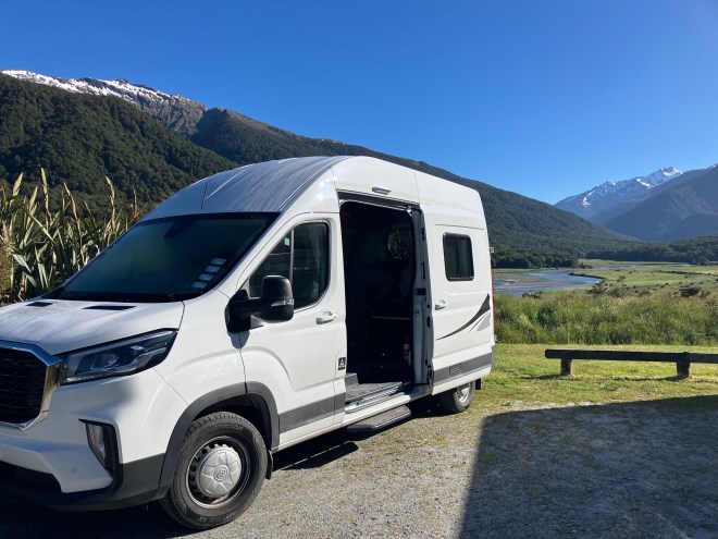 photo of a white camper van with the side sliding door open, backed into a campsite with a long green valley behind it and snow capped peaks in the distande