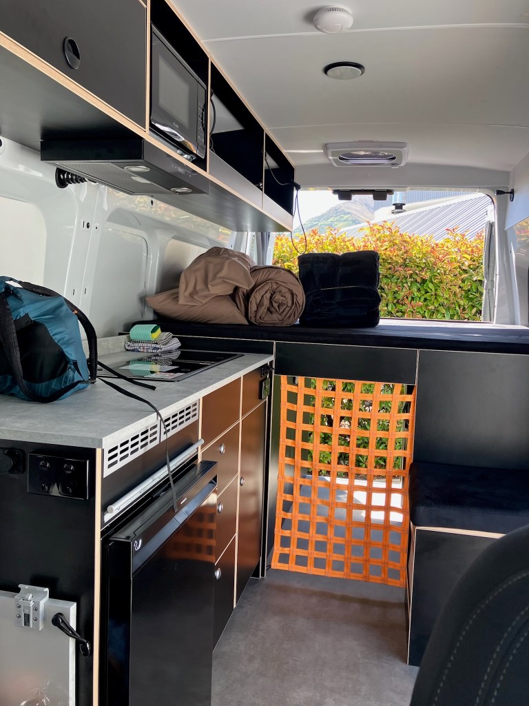 photo of the interior of a van with a small kitchenette on the left, cabinets above, a shelf in the middle, and a window in the back
