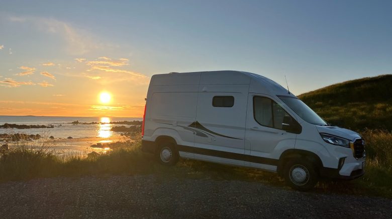 photo of a white campervan backed into an oceanside parking spot with a low orange sun behind it gleaming on the water
