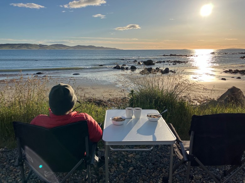photo of a person in a gray wool cap and red jacket sitting next to a white table bearing two coffee cups and cereal bowls, overlooking a beach with rocks, waves, and a low sun gleaming on the water.