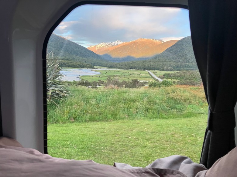 photo looking across a bed with brown covers through a window with curtain pulled pack. outside is a green-grass field, a long valley next to a blue river, with sunlit mountain peaks in the background