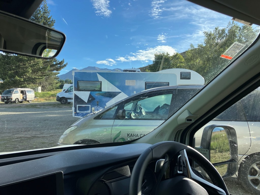 photo looking out a car windshield past the steering wheel at a row of cars, vans, and campervans