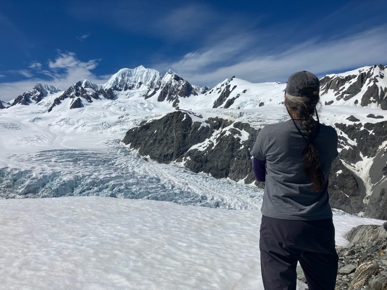 photo of a woman dressed in gray from the back, as she looks out across a rippled icy glacier field with rocky cliffs behind it