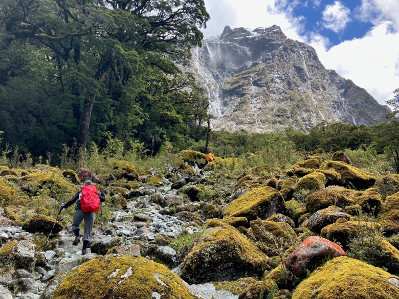 photo of a hiker wearing a red backpack climbing up a rock-strewn trail amid moss-covered boulders with a jagged peak lined with waterfalls in the distance