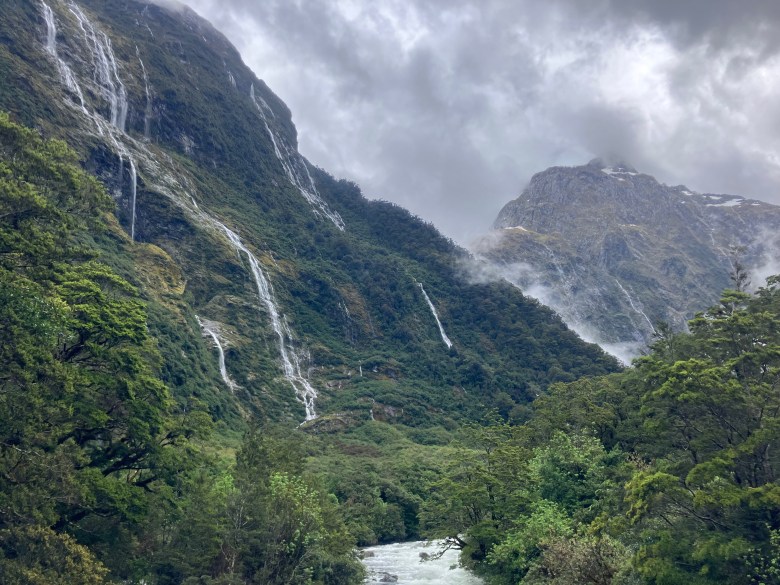 photo of two steep, green mountainsides lined with narrow white waterfalls and wispy white and gray clouds, with a river in the foreground