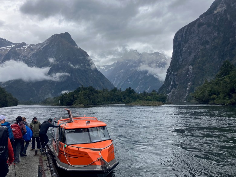 photo of hikers lined up to enter an orange, roofed boat on a wide expanse of dark blue water framed by steep rocky cliffs.