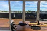photo looking across a brown wooden dining table through sliding glass doors onto a brown wooden deck, with treetops, a small mountain, and blue skies behind.