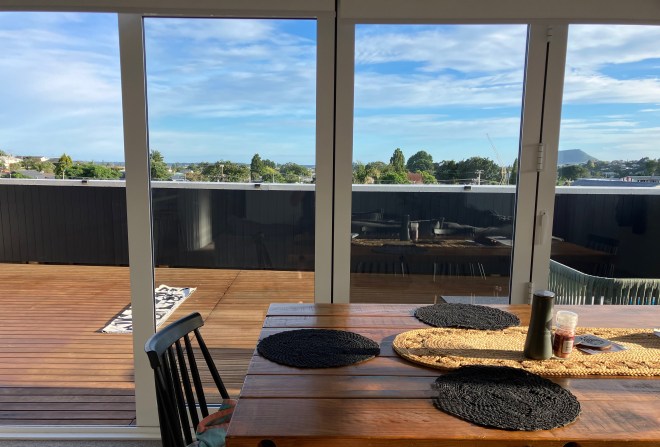 photo looking across a brown wooden dining table through sliding glass doors onto a brown wooden deck, with treetops, a small mountain, and blue skies behind.