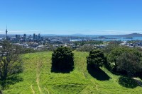 photo looking across a green grass-and-tree-covered hill to a cityscape with one tall spire and several high-rise buildings, with blue water and a green inverted cone-shaped hill in the background