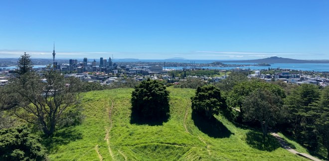 photo looking across a green grass-and-tree-covered hill to a cityscape with one tall spire and several high-rise buildings, with blue water and a green inverted cone-shaped hill in the background