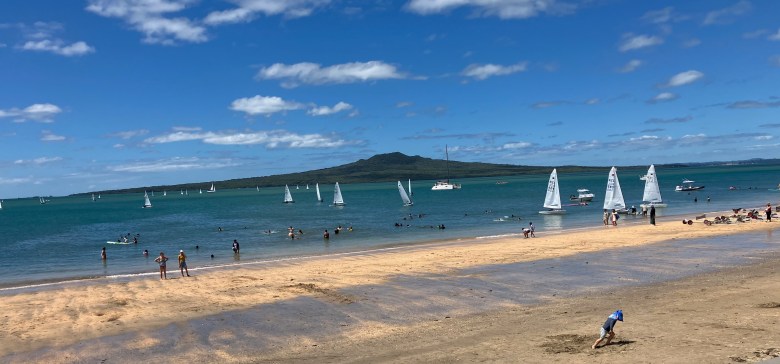 photo of a tan sandy beach with a little boy playing in the sand in the foreground, more people swimming and wading near the shore, dozens of small sailboats in the blue-green water, and an inverted cone-shaped green hill in the background.