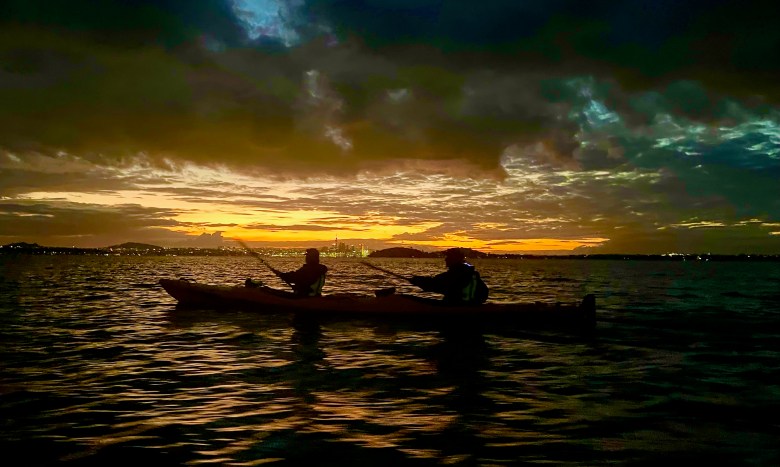photo of a silhouette of two people kayaking on a shimmering sea with an orange sunset under scattered clouds and the lights of a city in the distance