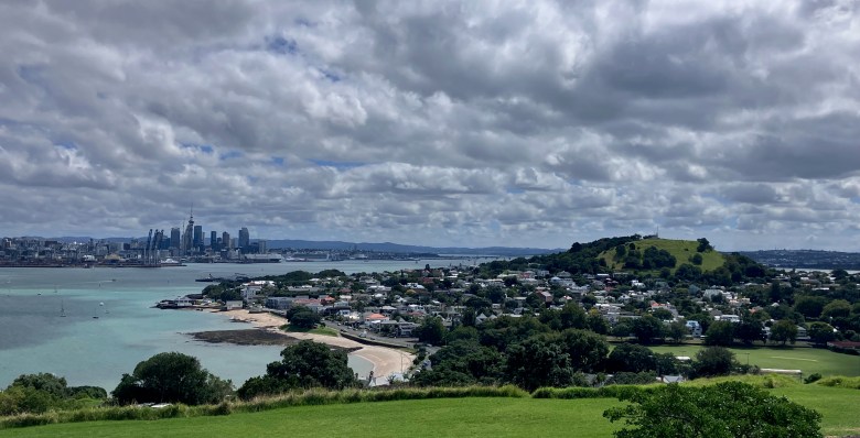photo of a golden sand beach curving along a green shore dotted by houses with green grass-covered hills in the foreground and background, and across the water lies a cityscape of tall buildings