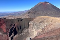 photo of a brown and red volcanic cone mountain with a white streak down its side, set against a deep blue sky, with a brown and sandy crater in the foreground that has a red and black jagged vertical hole ripped open in its site.