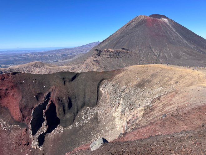 photo of a brown and red volcanic cone mountain with a white streak down its side, set against a deep blue sky, with a brown and sandy crater in the foreground that has a red and black jagged vertical hole ripped open in its site.