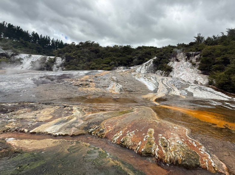 photo of brown, red, orange, rust, and white streams pouring across flat rocks with green trees and a cloudy sky behind