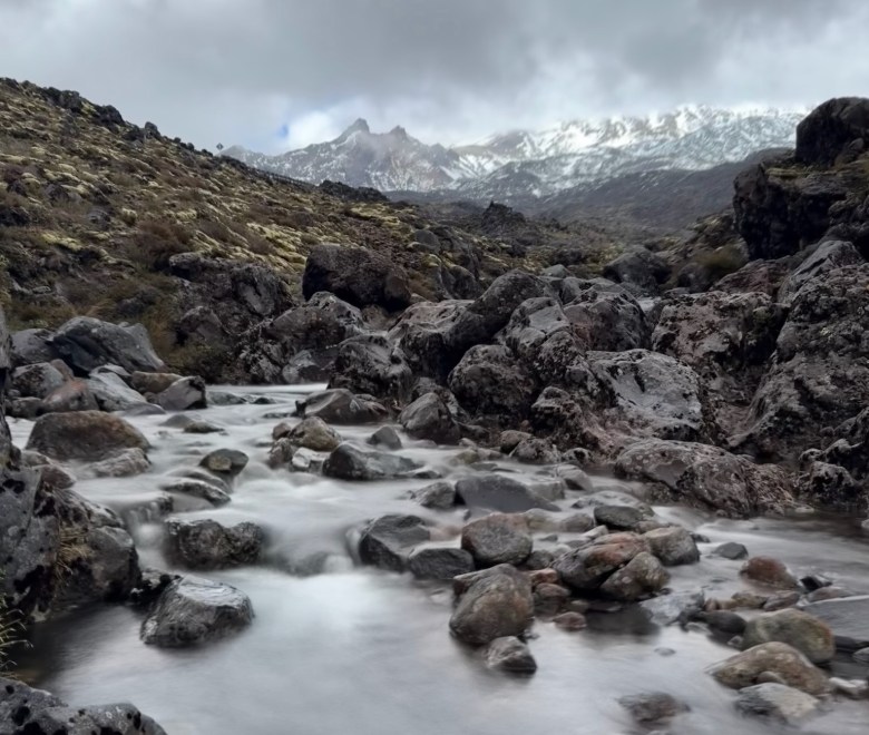 photo of a stream running through a tumble of rocks with a jagged, snowy mountain ridge under clouds in the background