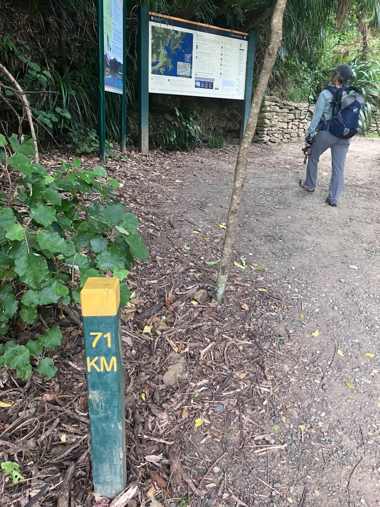 photo of a green and yellow trail marker with 71 KM on it, a gravel trail beside it, and a woman wearing a backpack up the trail looking at a trail map sign