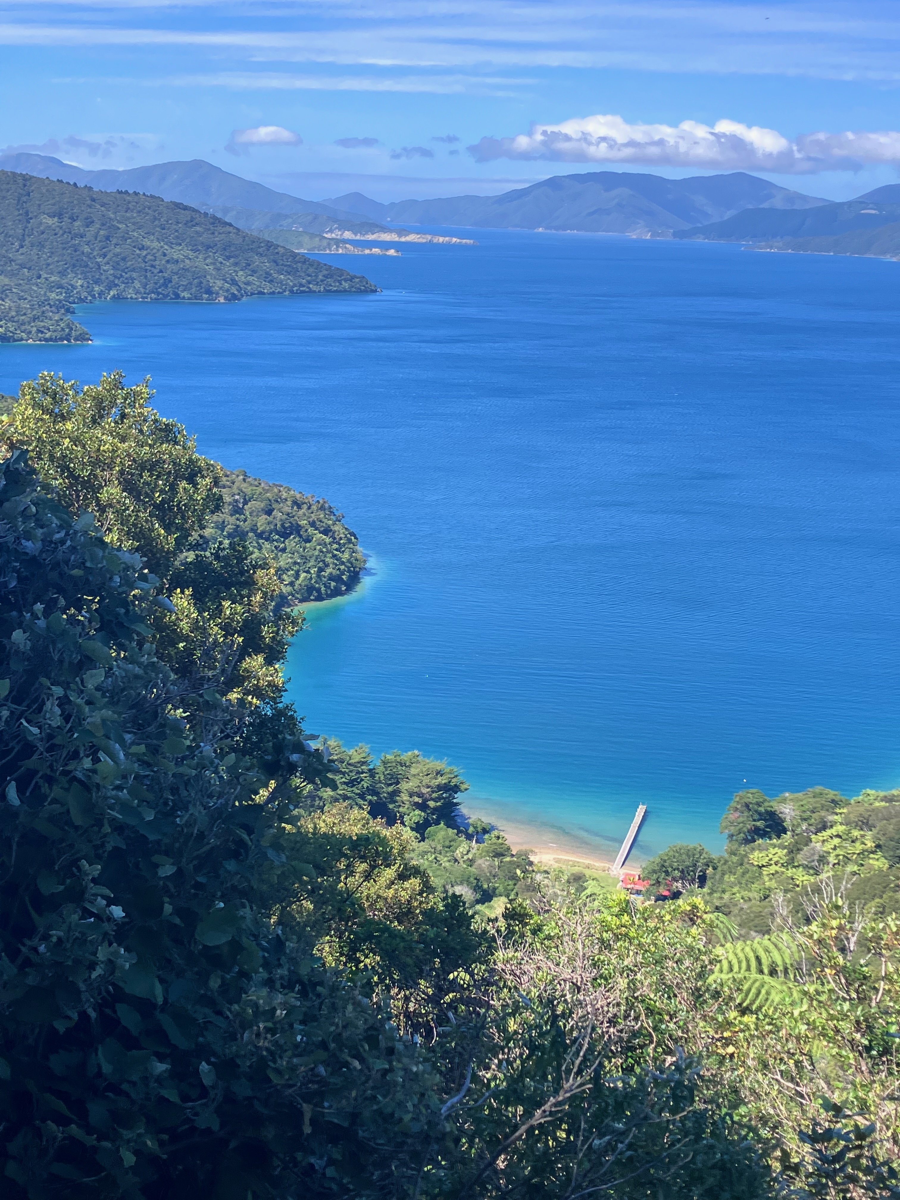photo from high up a hillside of a bright blue body of water ringed by green mountain slopes, with a narrow dock jutting into the water far below