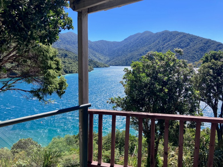 photo taken from a deck, with a wooden railing and post, overlooking a teal-blue sparkling body of water, green leafy trees, and green mountains beyond