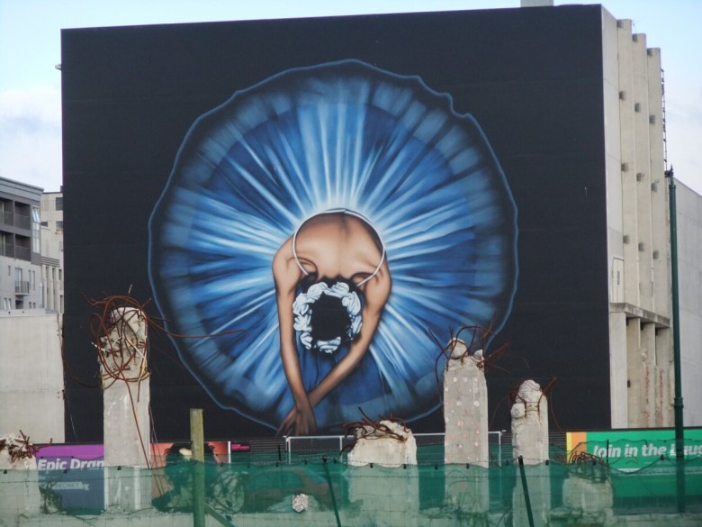 photo of a large mural on the side of a building of a ballerina from above with remnants of cement pillars in the foreground