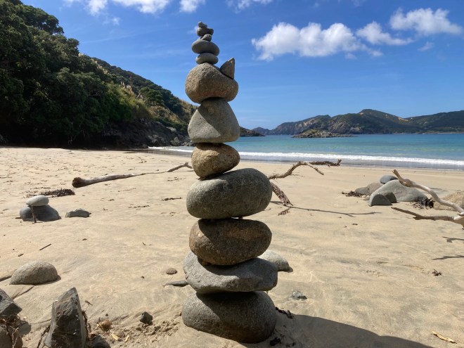 photo of a cairn of 12 rocks balanced precariously on the sand with blue-green water in the background