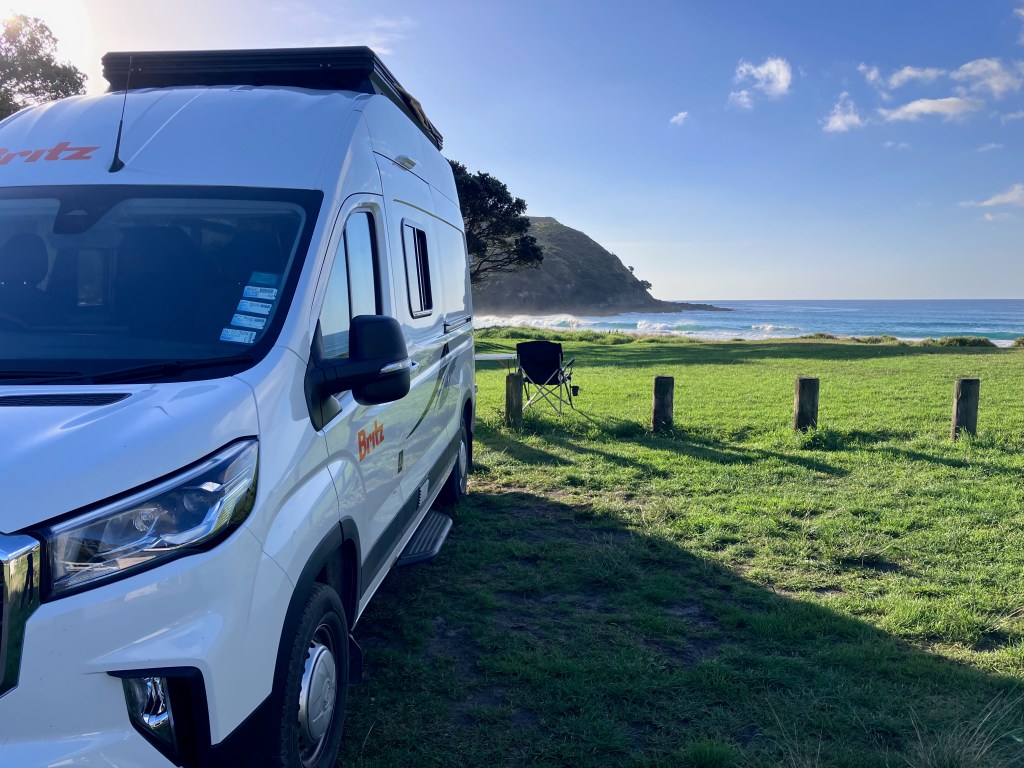 photo of a white van backed up on a grassy knoll with a black folding chair behind it overlooking the blue ocean