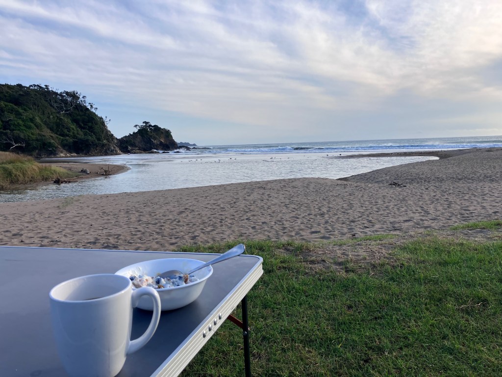 photo of a metal folding table with a white coffee mug, bowl and spoon on top, overlooking a sandy beach with water flowing in