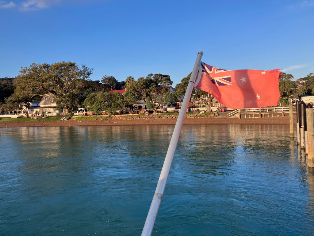 photo of a red New Zealand flag with the British Union Jack in the upper left corner, flying over a harbor with town buildings and a dock in the background