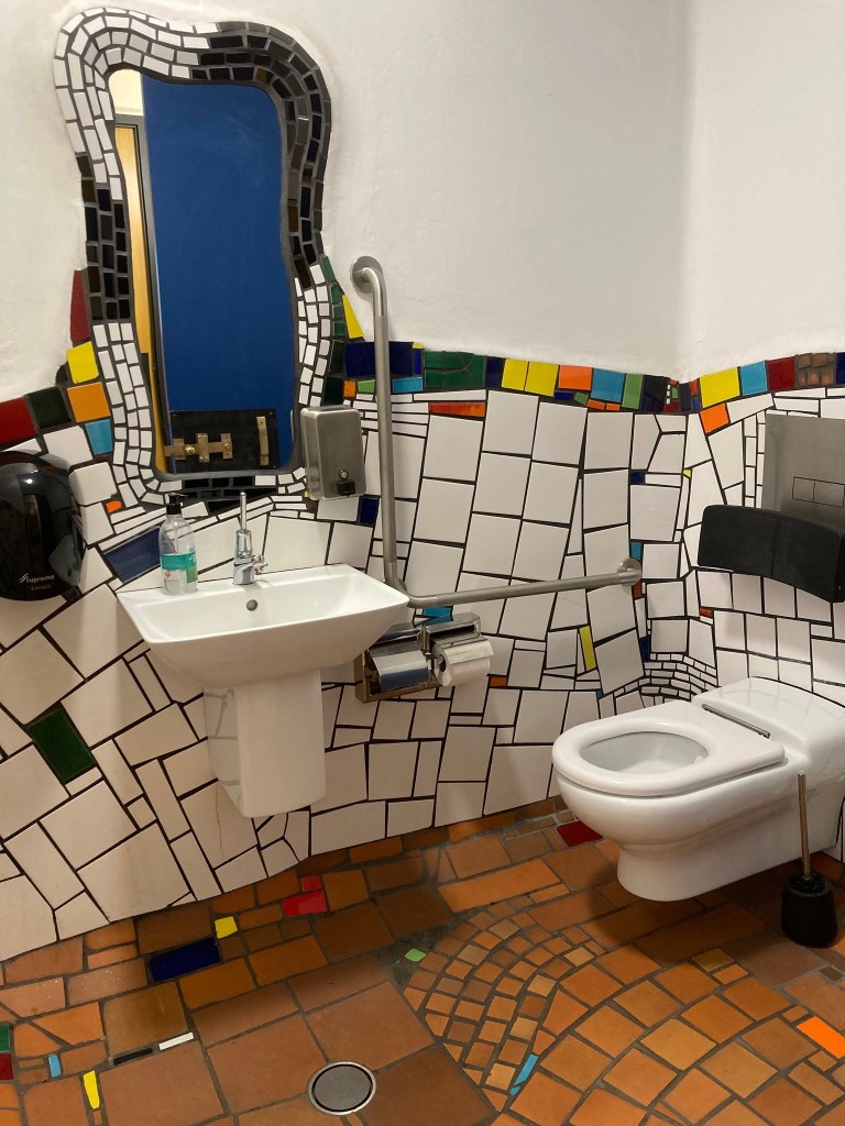 photo of a toilet, sink, and mirror in a misshapen, unevenly tiled, abstractly shaped room