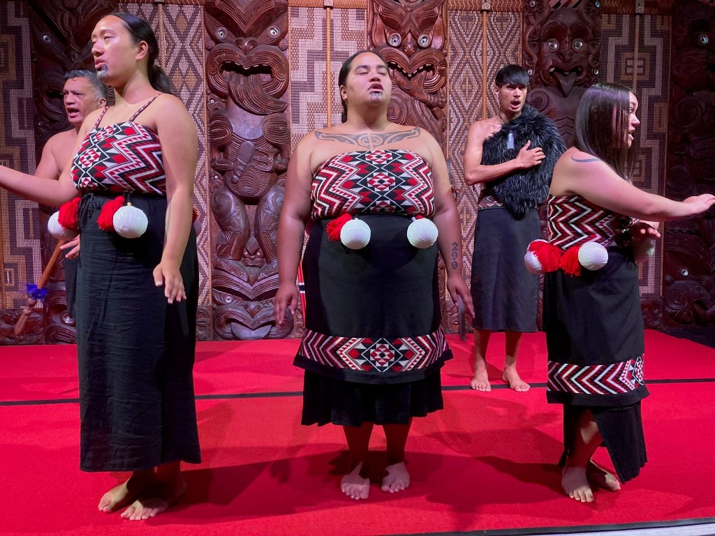 photo of three Maori women and two men in traditional dress performing a song. They wear black dresses with red, white, and black patterned tops.