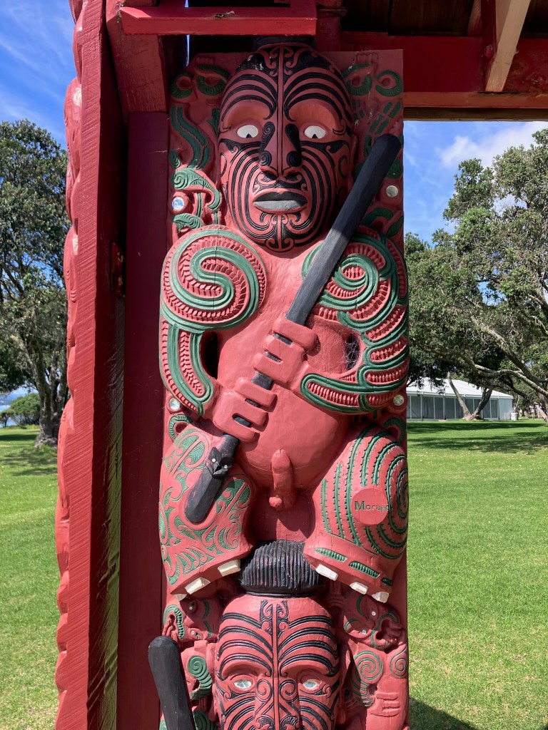 photo of a red Maori sculpture featuring a warrior with wide white eyes, holding a staff, with tattoos covering his arms and face