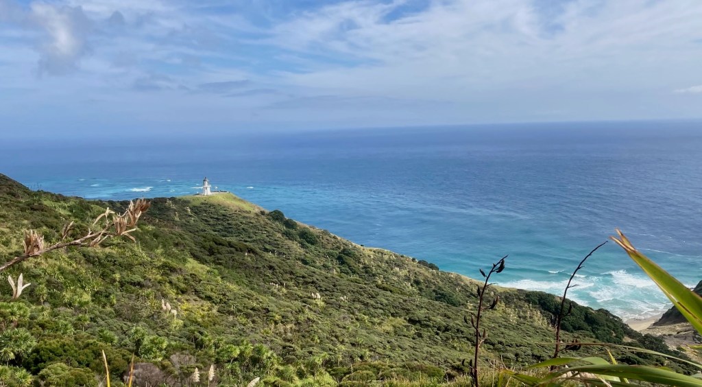 photo of a green hill with a small white lighthouse on its tip and green and blue water in the distance