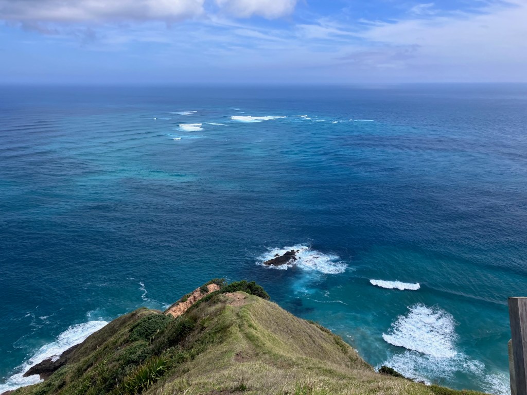 photo of a point of green land with white waves crashing on it and a swirling mix of blue and green water in the distance