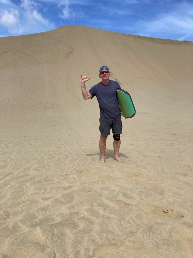 photo of a man wearing black t-shirt and shorts, sunglasses, and backwards baseball cap holding a green boogie board under one arm and flashing the Hawaiian "surfer" hand sign while standing in front of a tall sand dune