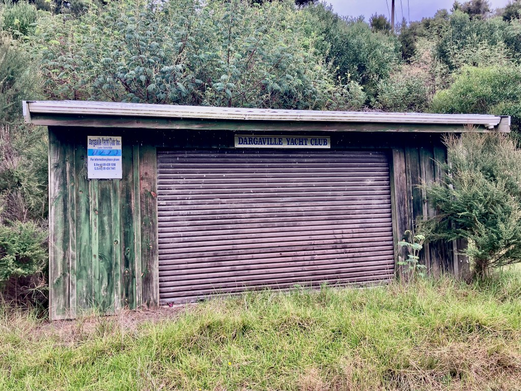 photo of a small brown shed with grass and bushes around it and a sign saying "Dargaville Yacht Club"