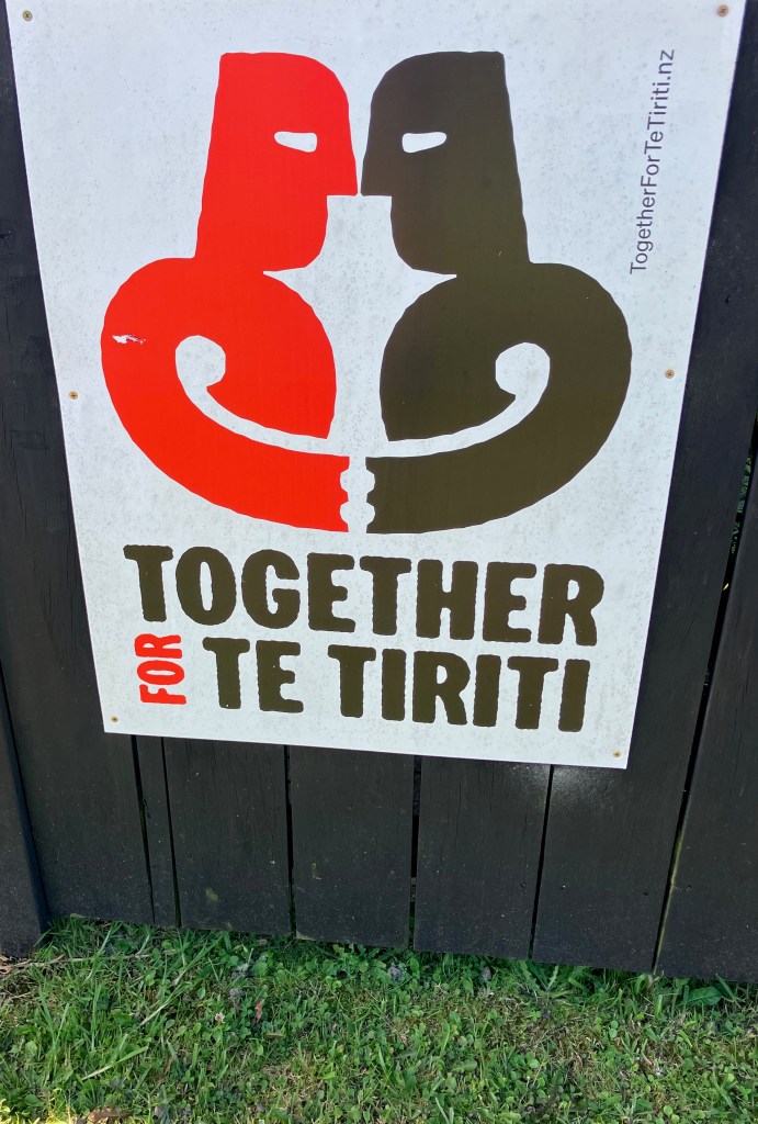 photo of a sign with two emblematic people, one black and one red, facing each other and holding hands, with the words below "Together for Te Tiriti"