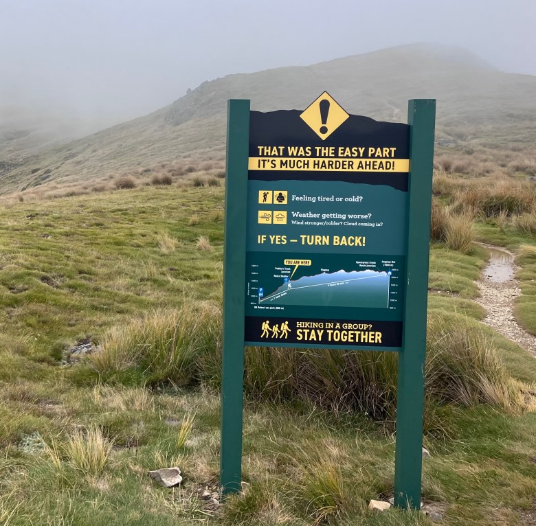photo of a green and yellow sign next to a hilltop hiking path. The sign has a big exclamation point and a headline THAT WAS THE EASY PART. IT'S MUCH HARDER AHEAD!