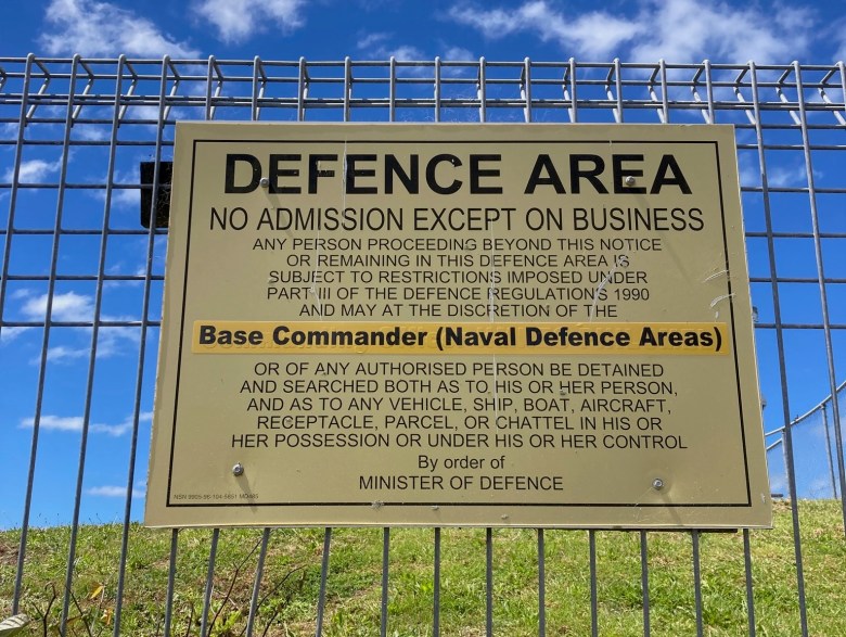 photo of a gold sign with black letters spelling "DEFENCE AREA", attached to a wire fence.