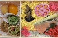 photo looking down on a light brown wood box with orange fruit, a green fiddlehead, and two undefinable mushy things on the left, then yellow shreds, a few brown mushrooms, a pink slice of something with holes, a piece of salmon, and assorted other colorful items on rice